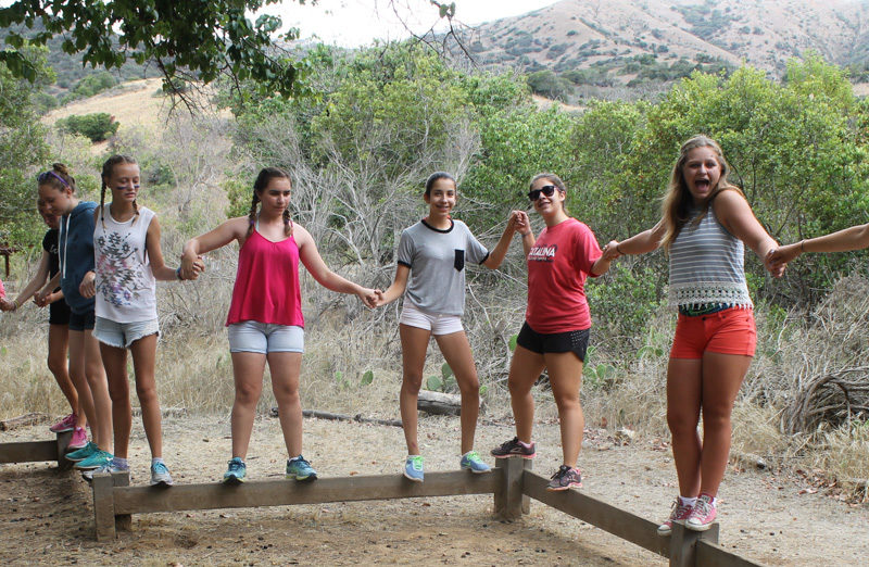 girls attempting to balance on a balance beam and form a human chain by holding hands
