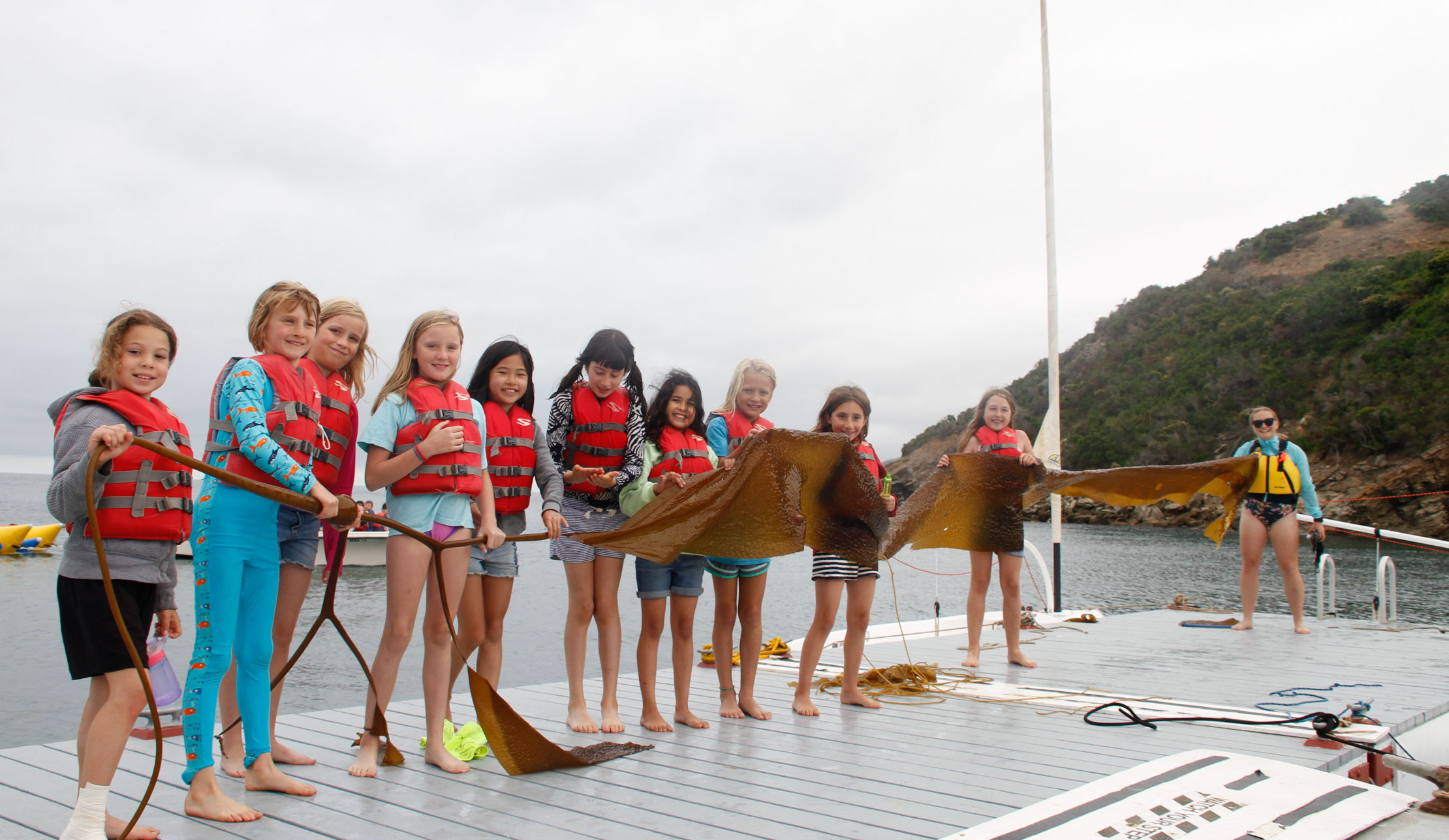 girls on the dock wearing lifejackets with a counselor holding a very long piece of kelp